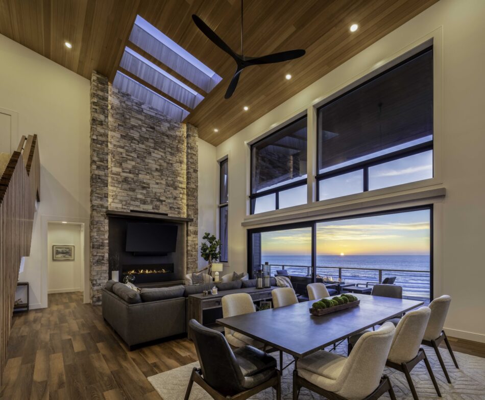 A dining room with huge windows overlooking the ocean. A dining table is in the foreground and a large, lit stone fireplace and a large leather couch sit in the background.