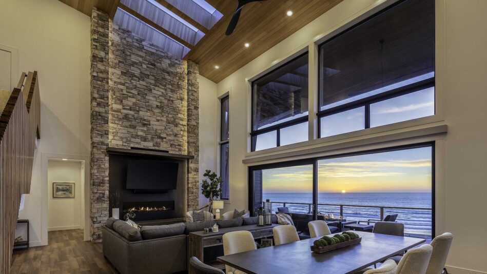 A dining room with huge windows overlooking the ocean. A dining table is in the foreground and a large, lit stone fireplace and a large leather couch sit in the background.