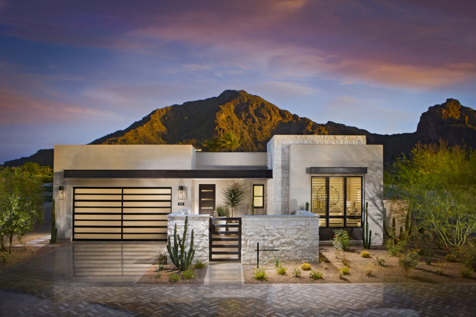 Rectangular modern home is built of white walls and white stone veneer surrounded by desert vegetation and a stone mountain in the background.