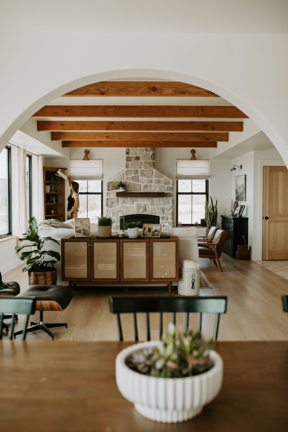 The view of a living space through an arched doorway. The living space has exposed beam ceilings, an exposed stone fireplace and tan hardwood flooring.