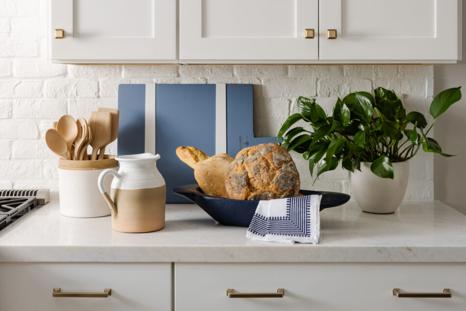 This kitchen features a white brick veneer backsplash. There is a jar of kitchen utensils, a potted plant, a bowl of bread, a blue cutting board and a ceramic pitcher on the counter.