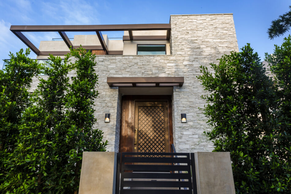 A modern home with light gray stone veneer siding and a unique wooden front door. The home has a metal gate with large trees on either side of the gate.