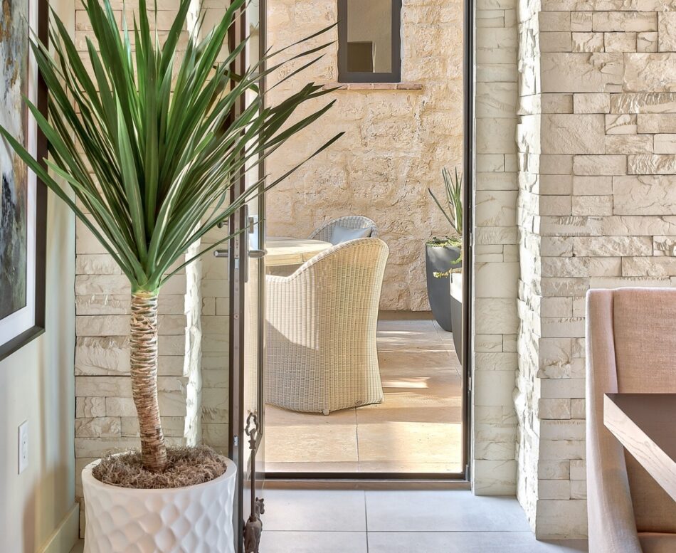 A palm tree is in the foreground and an iron doorway opens up to show a table with wicker chairs in the next room. All the walls are made from stone.