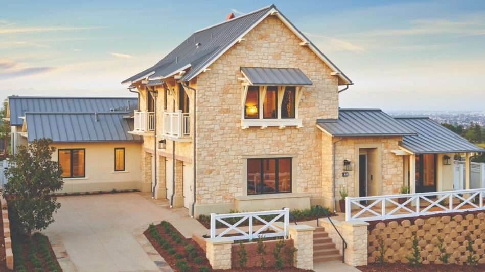 A tan, stone home sitting on the top of a hill. There is a white fence and landscaping surrounding the house, and a sidewalk in front of the house.