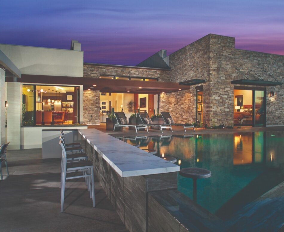 Large patio area with raised pool with seating area on the edge and a large home with stone veneer finished siding.