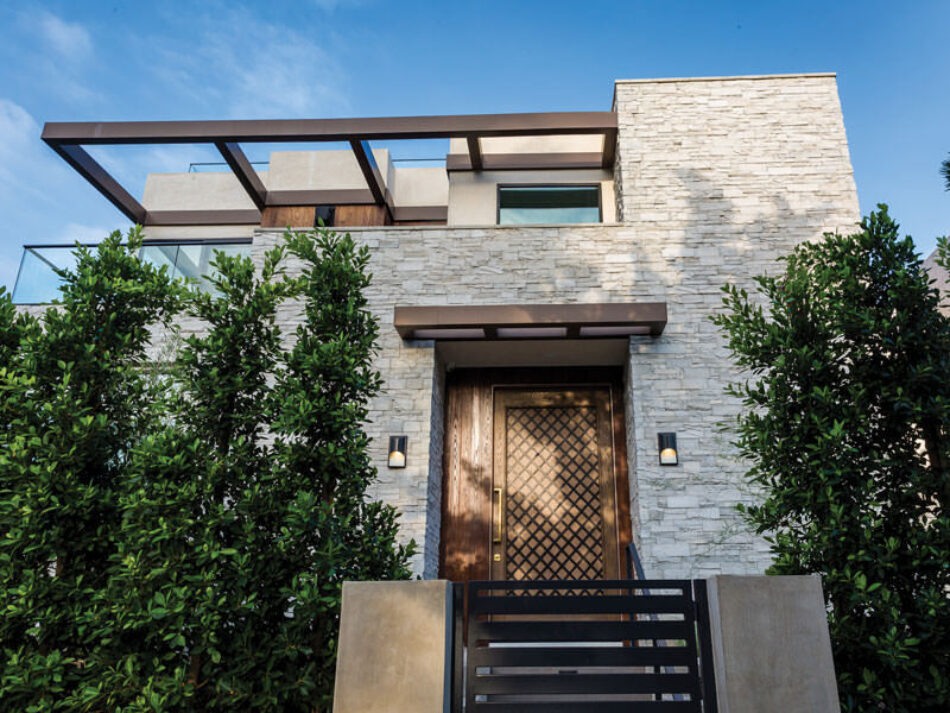 An image of the front of a contemporary home. The home is stone and gray, and large trees are on either side of the front door.