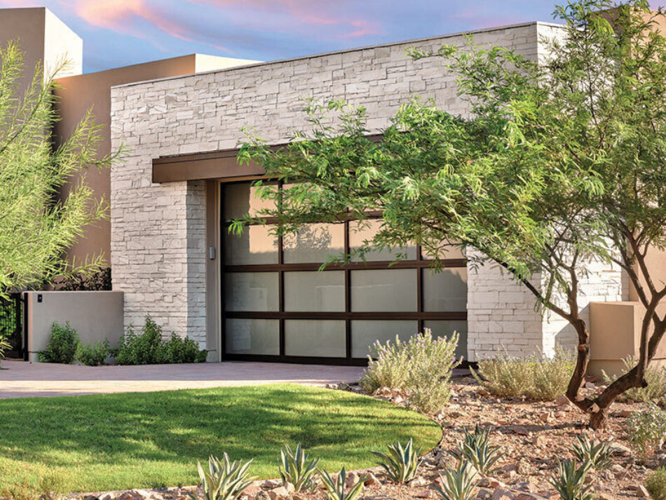 This home features a garage with stone veneer siding. There are trees incorporated into the home’s landscaping, along with other green plants.