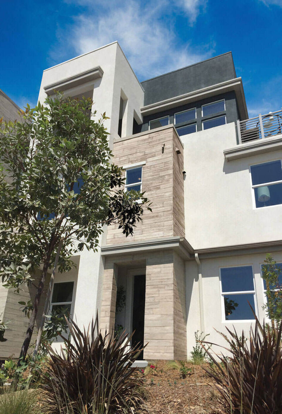 The exterior of a three-story modern home made of concrete and wood veneers. There are plants and trees in front of the house.