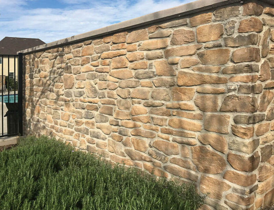 A large and tall stone wall with grass in front of it, and a metal gate at the end of it.