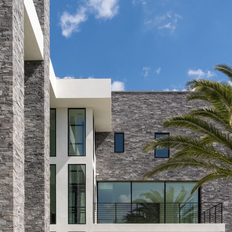 A modern custom home features an extensive use of cut ledge stone, contrasting smooth white plaster and oversized black framed windows.