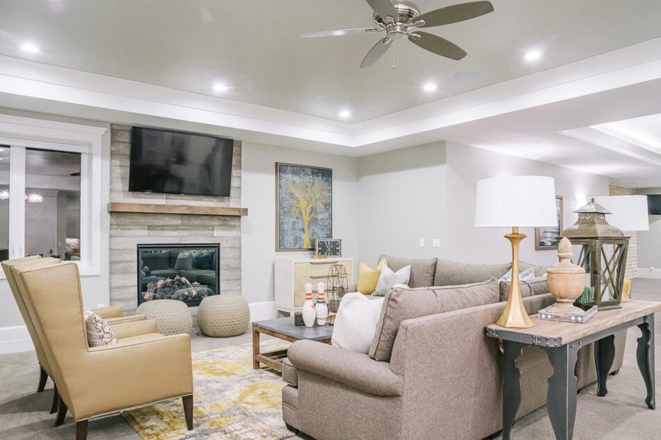 A bright living room with a sectional couch and two chairs. There is a gas fireplace with a TV mounted above it, on a wood veneer wall.